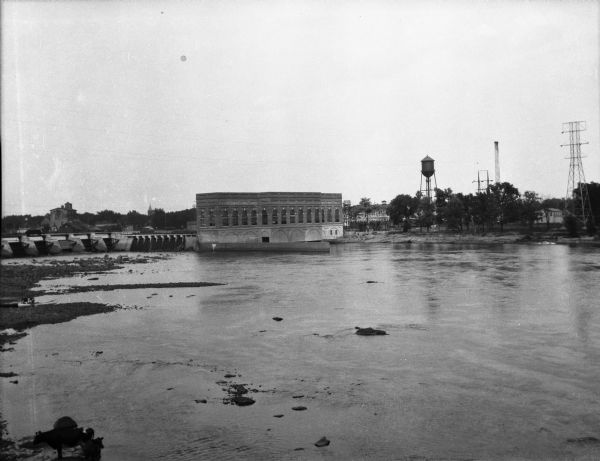 Exterior of the Consolidated Water Power and Paper Company, built in 1918. The view from across the Wisconsin River shows the company building, a dam, as well as the city's water tower. On the bottom left, cows stand along the shoreline.