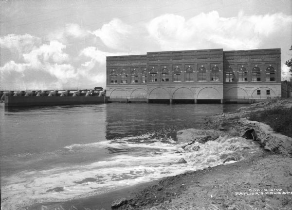 Exterior of the Consolidated Water Power and Paper Company, built in 1918. The view from a bank of the Wisconsin River shows the company building and dam.