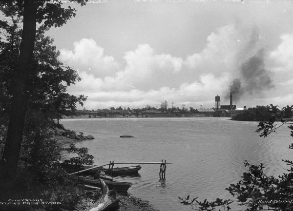 View taken from the Paper Mill Road toward the Wisconsin River Pulp and Paper Company paper mill. On the bank in the foreground are small boats. The paper mill's water tower and smokestack stand in the distance. The mill was later purchased by Consolidated Papers, Inc. before eventually closing.