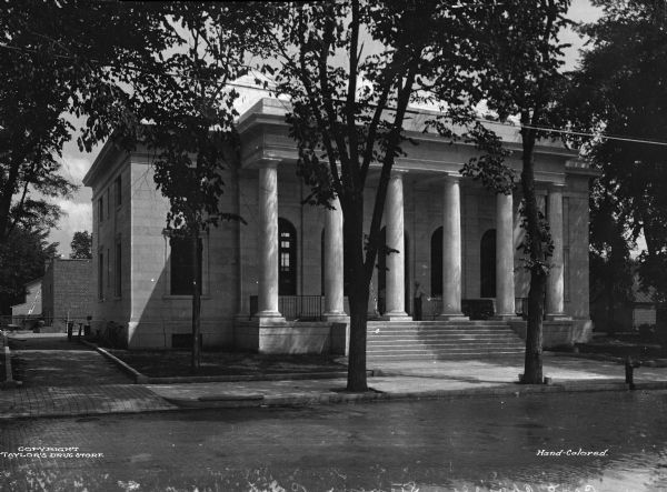 Exterior view of the Post Office, built in 1919. The view from across Main Street shows men standing at the main entrance of a stone building built in the classical revival style. Stairs lead to the front doors beyond a row of Doric columns. Bicycles lean against the left side of the building.