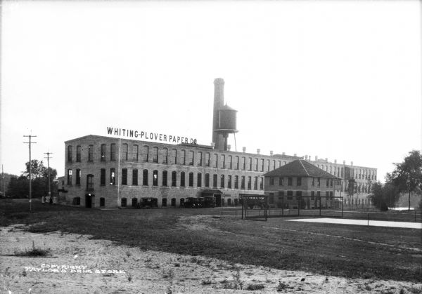Exterior view of Whiting-Plover Paper Company, founded in 1892. The view features a three-story brick building with automobiles parked in front.