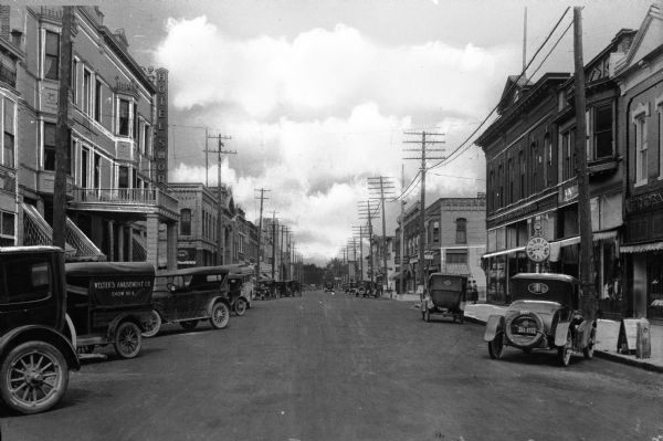 View down Main Street. Automobiles are parked along the curbs, pedestrians are on the sidewalks in front of storefronts. Hotel Swoboda can be seen at left; the hotel opened in 1901 and was renamed in 1917.  On the right stands the L. M. Washburn Company, which opened in 1870. An automobile on the left has a sign that reads: "Welter's Amusement Co. Show No 4".
