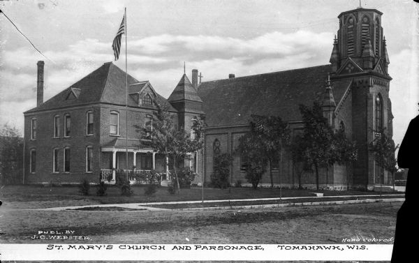 Exterior of Saint Mary's Church and Parsonage. The view from across the street shows a United States Flag in front of the parsonage. Caption reads: "St. Mary's Church and Parsonage. Tomahawk, Wis."