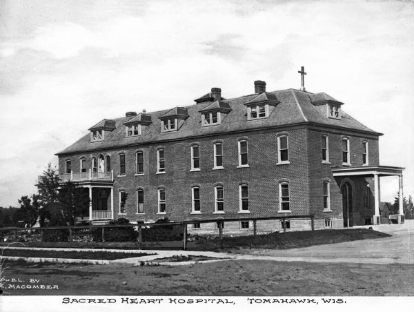 Exterior of Sacred Heart Hospital. The brick hospital building features a porch, balcony, and a covered entrance. Published by C. E. Macomber.