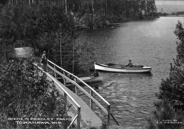 View of Bradley Park, named in 1910 after the consolidation of 105 acres previously referred to as Prospect Point and Hog's Back. A man observes men boating from a wooden walking bridge and a dirt path continues into the woods. Published by J. C. Webster.