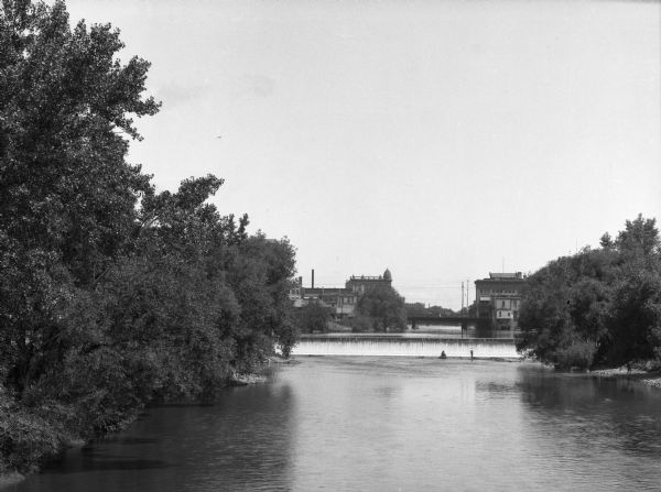 View of the Rock River. Two men stand before a dam and, beyond them, a woman stands on a bridge. Downtown buildings are visible in the background.