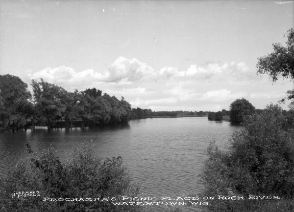View of Prochazka's picnic place on Rock River. The view features a small boat docked on the left near trees which crowd the shoreline. Caption reads: "Prochazka's Picnic Place on Rock River, Watertown, Wis."