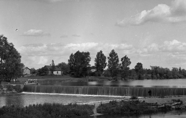 View of the Rough and Ready Dam, a concrete electrical dam built in 1907. Men accompany a group of boys on the far bank of the river. One boy swims near the dam, while another on the right wades in the water by the dam.