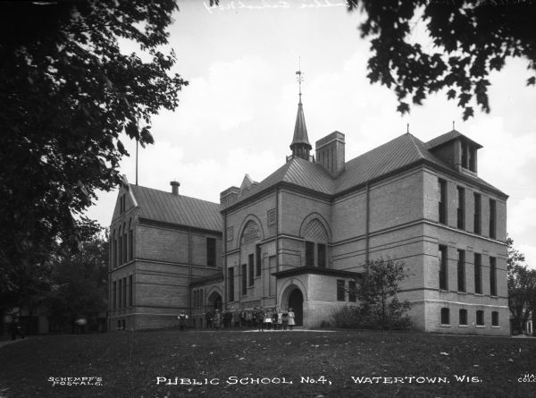 Exterior of Public School No. 4, built in 1883. The brick building entrance has a sign that reads: "High School," and features Gothic arches, long, rectangular windows, and a central cupola. Young children stand outside the main entrance. Caption reads: "Public School No. 4, Watertown, Wis."