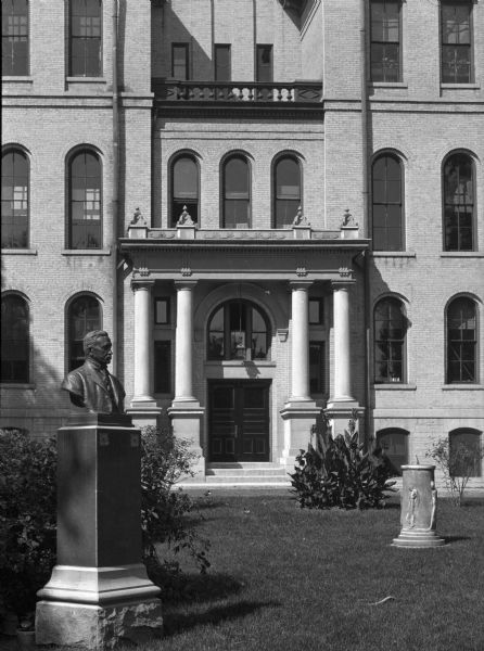 Exterior of "Old Main" at the State Normal School, founded in 1868.  The view features a main entrance flanked by columns. A statue and a sundial stand on the lawn before the entrance.