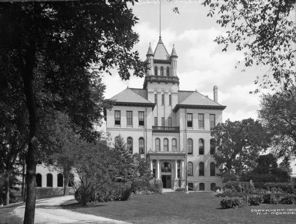 Exterior of "Old Main" at the State Normal School, founded in 1868. A dirt road leads to the main entrance, flanked by columns. A central tower is above the entrance.
