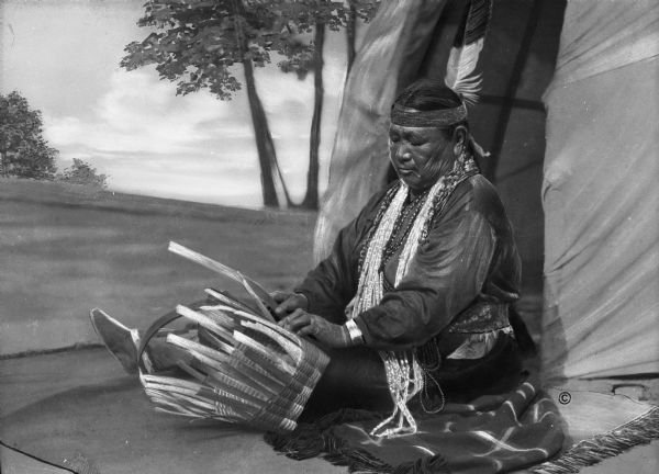 Mary (Stacy) Yellowthunder, (Cowboy Woman) weaving a basket while seated on a blanket. A dwelling and trees can be seen in the background. She is the daughter of Henry Stacy of Wittenburg, Wisconsin.