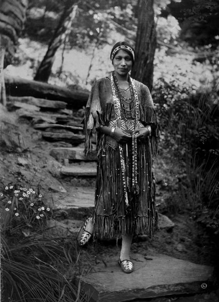 View of a Winnebago (Ho-Chunk) Indian woman in traditional dress. The woman stands on stone steps, surrounded by flowers and trees.