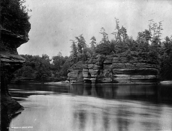 View of Steamboat Rock, a rock formation on the Wisconsin River. A small boat can be seen on the shore of the river. Caption reads: "Camera Work by Sweet MPLS."