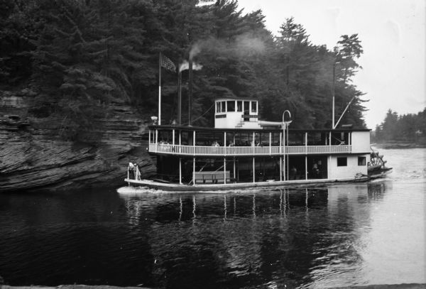 View of the "Winnebago," a paddle-wheel steamboat on the Wisconsin River. Passengers sit on the top deck and a man and two women look over the bow.