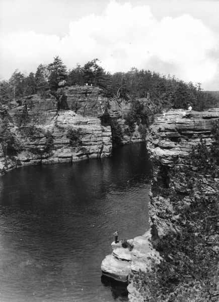 Elevated view of a man standing on a rock formation at the base of Romance Cliff and High Rock while a woman sits on the ledge above, looking out over the Wisconsin River.