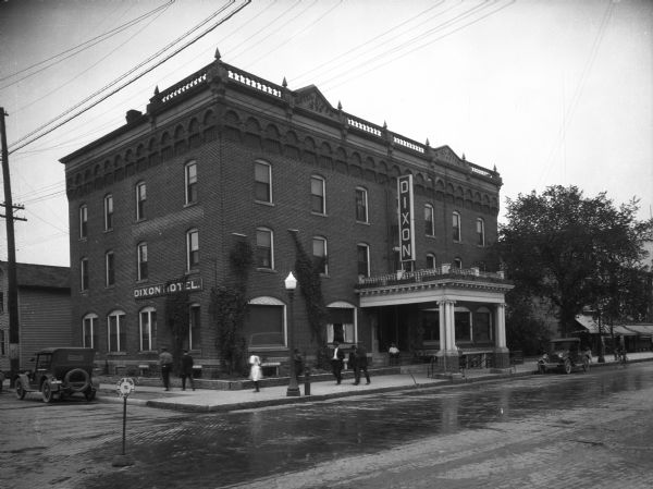 Exterior view across street towards the Dixon Hotel, featuring an overhang over the entrance supported by groups of Ionic columns. There is a balustrade on the roof along the front and part of the sides of the hotel. Pedestrians are walking along the sidewalk around the hotel. Signs around the building's balustrade read "1808" and "1918." In the street, another sign in the intersection reads: "Slow Down & Keep Right."