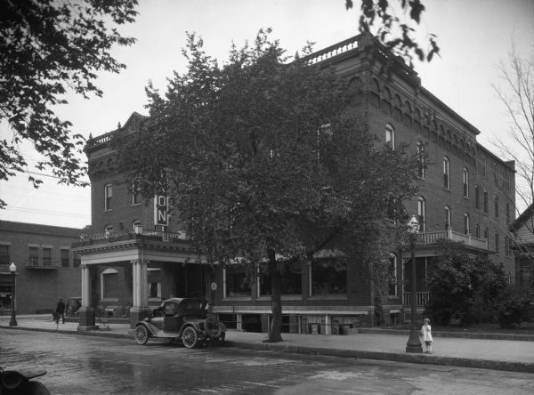 Exterior of Dixon Hotel featuring an overhang supported by groups of Ionic columns. Pedestrians walk along the sidewalk around the hotel, and a child stands by a lamppost. A car is parked in front of the main entrance.