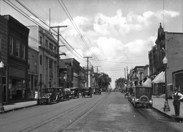 Automobiles drive past storefronts and can be seen parked on either side of Grand Avenue. Pedestrians walk on the sidewalk near the Citizens National Bank on the left. Further in the distance, large columns mark the entrance of the Wood County National Bank, built in 1911. A man stands on the right at the edge of the curb.