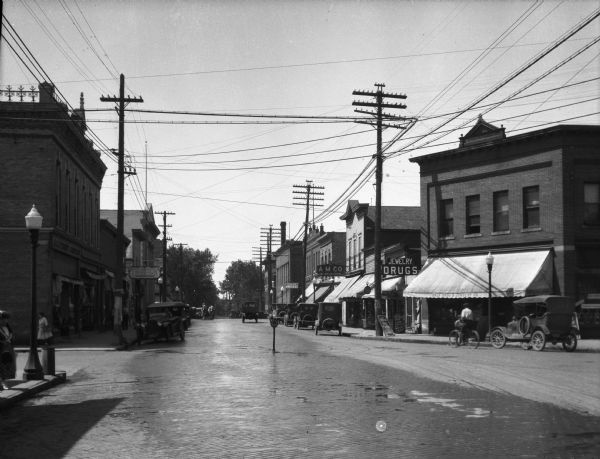 View of the business section featuring jewelry, shoe, and drug stores and other storefronts. Women and men walk down the sidewalk near cars parked on the cobblestone street. A man steers horses and a carriage toward an intersection where a sign reads, "Keep to the Right."