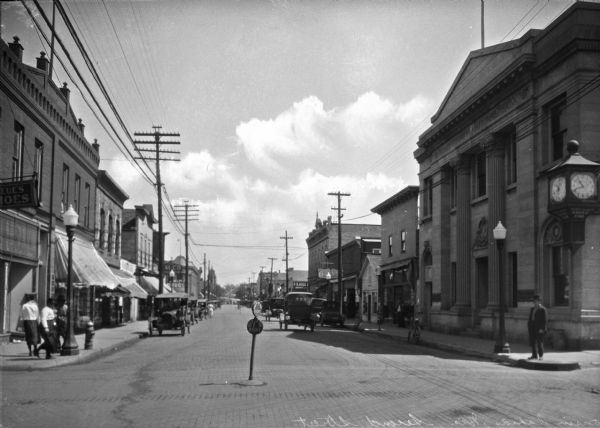 View down Second Street. Automobiles are parked on either side of the street near bicycles and pedestrians. Stores line the street and the Wood County National Bank stands on the corner at right.