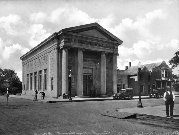 Exterior of the First National Bank, built in 1919. Men stand outside the building, one drinking from a fountain. A man stands across the street near the curb.