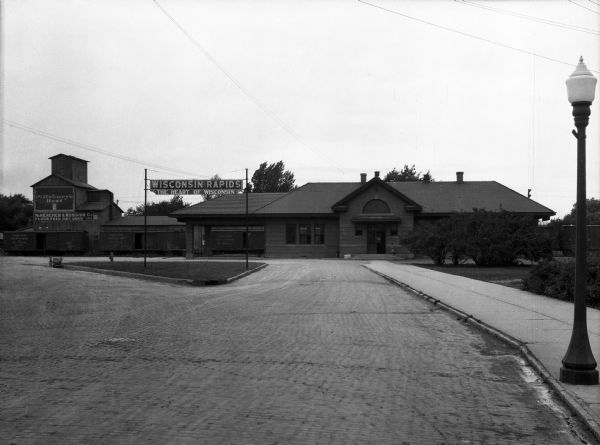 View down cobblestone street toward a girl standing on the steps of the front entrance of a railroad station. Railroad cars on the left of the station are labeled Chicago, Milwaukee, and St. Paul Railroad, a Class I railroad that began operation in 1847. A sign before the station reads, "Wisconsin Rapids, The Heart of Wisconsin." To the left is a factory with a sign that reads: "Pillsbury's Best, McKercher & Rossier Co. Flour, Feed, Hay, Grain, etc."