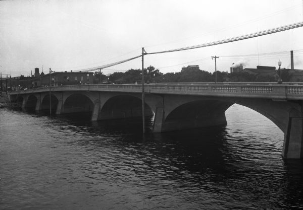Elevated view over water toward a horse and carriage and pedestrians crossing the Grand Avenue Bridge, built in 1922. Smokestacks and other buildings are on the far shoreline.