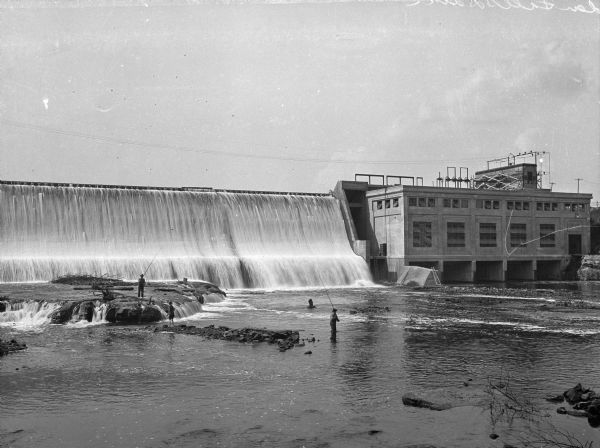 View across water toward the Cedar Falls Dam, built in 1910. Men are standing in the river and fishing at the base of the dam.