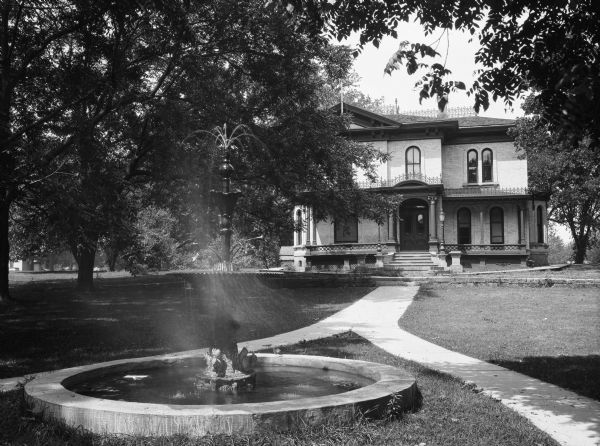 View of an elaborate fountain decorated with squirrel figurines at its base. A path leads to a two-story brick house beyond the fountain. Lampposts and columns flank the main entrance.