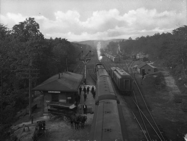 Elevated view of a railroad station at Beaver Lake. Men gather in front of the station on the left where horses and carriages are parked. Signs on the station read: "Stag Tobacco" and "Western Union Telegraph and Cable Office." Hills are in the distance.