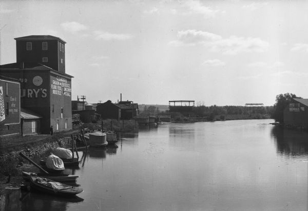 View of the Fox River. Small boats are docked along the left bank where factory buildings stand. The facade features advertisements for Wrigley's Spearmint Chewing Gum, stating "Chew it after every meal."  The next building carries a sign reading "C.S. Morris, Grain and Produce, Flour, Feed, Lime, Cement, Brick Tile, and Coal." A man stands outside the factory building near the river. On the right bank, a lumber company stands on the edge of the water.