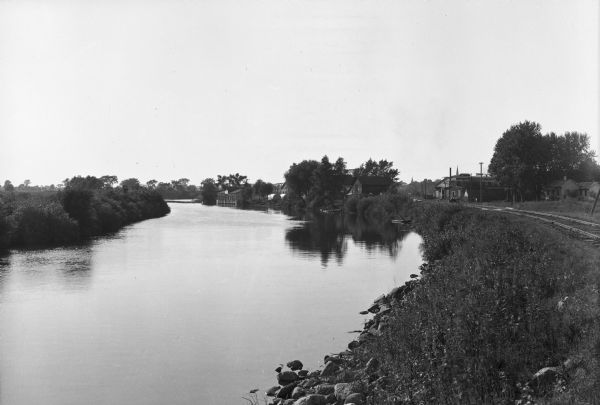 View along bank of the Fox River. A railroad track on the right bank extends toward buildings of the city.