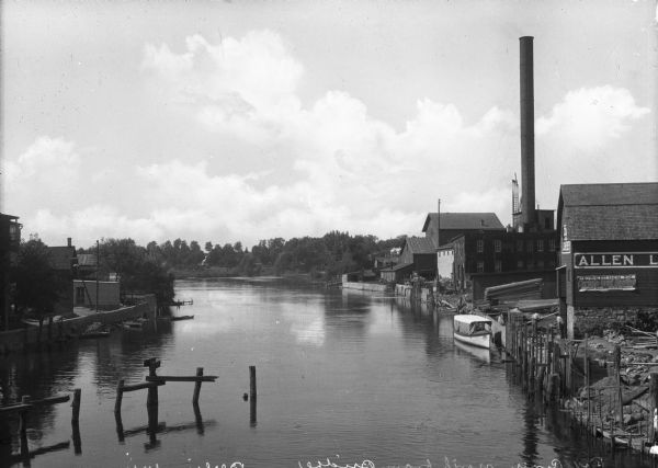 View of the Fox River, taken from a bridge looking north. Allen Lumber Company, founded in 1888, stands on the right bank. A sign on the building states, "We Sell the Best Roofing Made, Certain-teed Roofing, Guaranteed 15 Years - In Rolls and Shingles." Men work near the river's edge where a boat is docked.