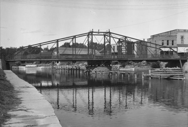 View of a bridge on the Fox River. A sign on a factory building on the right states: "Farm Implements, Wagons, Carriages, Etc." Further in the distance is the Allen Lumber Company, founded in 1888.