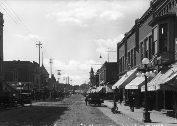 Westward view of Sturton Street, a dirt road lined with storefronts.  Automobiles and horse-drawn carriages are parked along the street. On the right a man stands near his bicycle, while other men walk down the sidewalk and lean against shop windows.