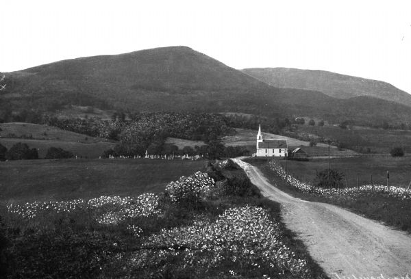 View down a dirt road leading to Mount Equinox in the background.   A church is in the valley near the road, and a cemetery is located on the other side of the road.