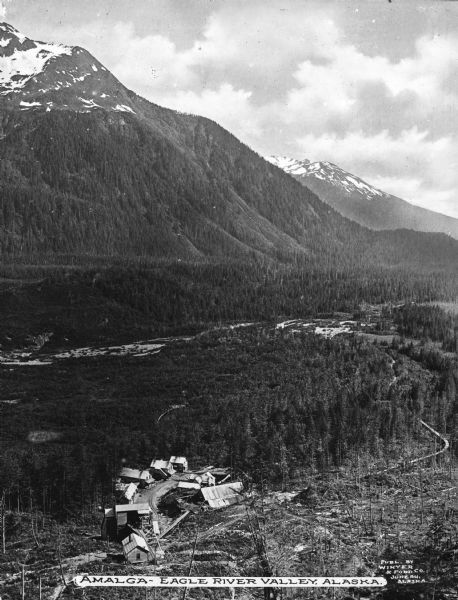 Elevated view of a small group of homes standing among trees at the base of a mountain in Eagle River Valley. Caption reads: "Amalga - Eagle River Valley, Alaska."