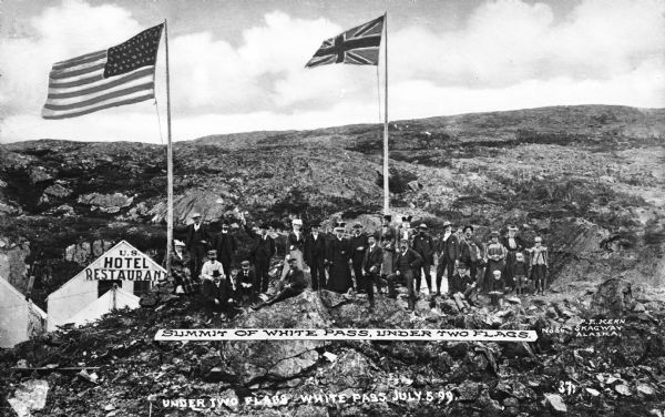 View of a tourist group at White Pass, a mountain pass located between Alaska and Canada. A shack has a sign that reads: "U.S. Hotel Restaurant" is to the left of the group. The United States and Canadian flags are flying on flagpoles. Caption reads: "Summit of White Pass, Under Two Flags." Another caption below reads: "Under Two Flags, White Pass, July 5, '99."