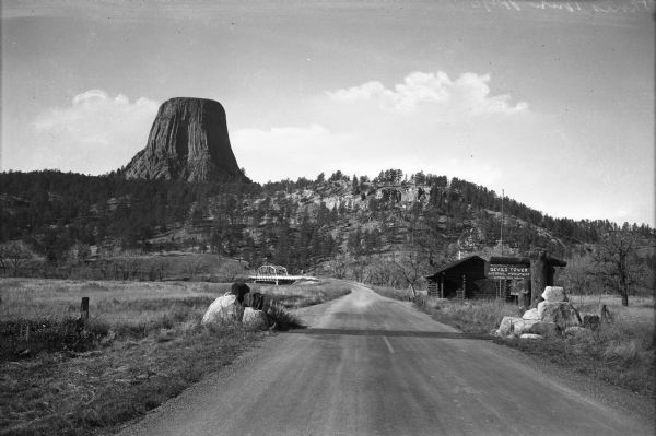 View of a highway leading towards Devils Tower. A sign before a cabin on the right reads, "U.S. Department of Interior, Devils Tower, National Monument, National Park Service."