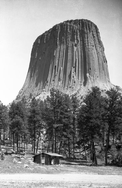 View of the distant Devils Tower, a large striated rock formation. A couple stands in the foreground near a small log cabin among trees.