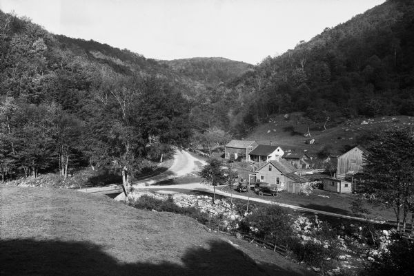 View of a mountain pass called Dunville Notch. A group of farmhouses are located among trees and a dirt road. Men gather near parked cars and a sign near them reads, "Beech Nut Chewing Tobacco, Famous for Quality."