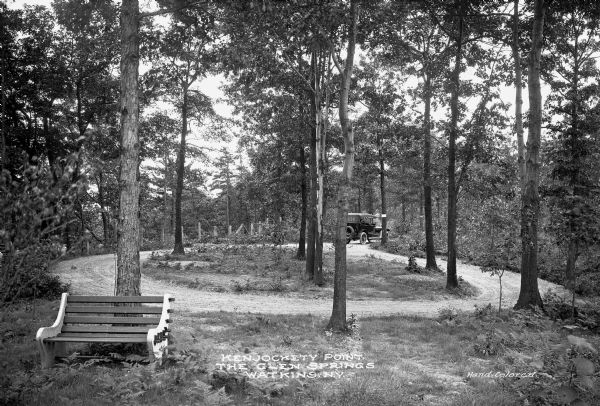 View of Kenjockety Point at Glen Springs. A bench and automobile are in the small wayside in the woods. Caption reads: "Kenjockety Point, The Glen Springs, Watkins, N.Y."