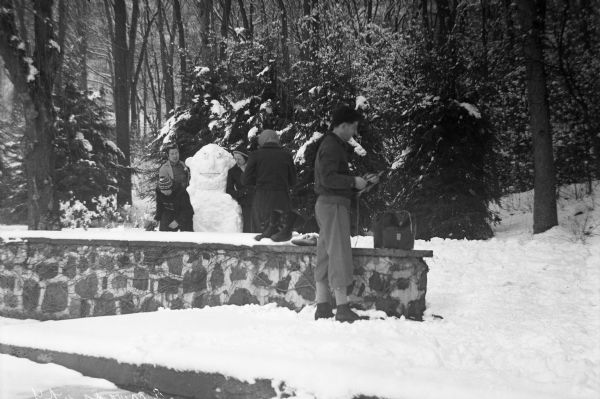 A group of people build a snowman while standing near a stone wall which has boots placed upon it. A wooded area is in the background.