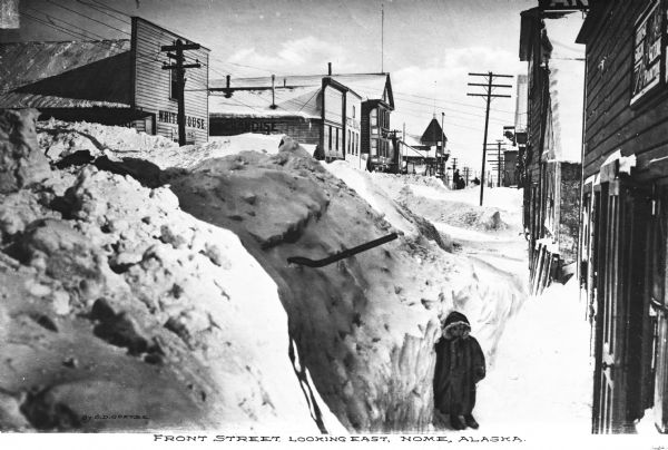 View of Front Street, looking east.  The street is surrounded by stores and other buildings and covered with snowdrifts.  A child wearing a parka stands on the right.