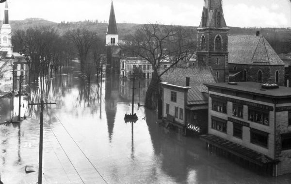 Elevated view of a flood scene looking up Main Street showing Kellog-Hubbard Library, the stone building which opened in 1895, and churches. Men and women look out the windows of a building with a sign that reads, "Montpelier Crackers, Established 1828, C.H. Cross & Son."