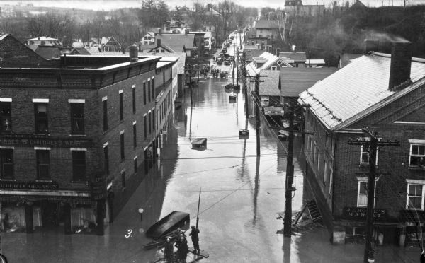Elevated view of a flood scene showing East State Street with people on a wooden makeshift raft investigating. They are floating past stores and markets. In the distance automobiles are parked on the higher portion of the road.
