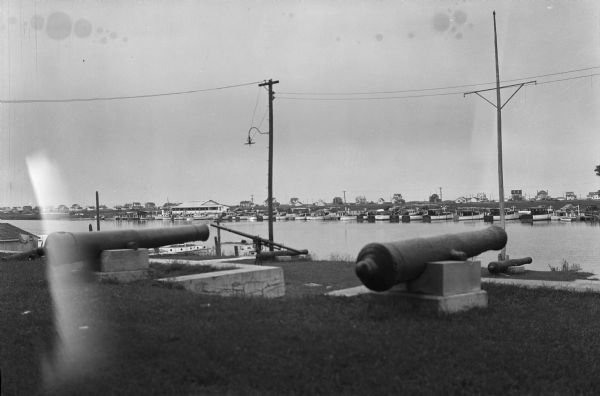 View of cannons mounted on a shoreline. Docked boats are across the river.  