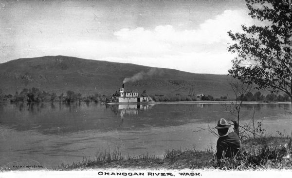View of a steamboat in the Okanogan River with mountains on the opposite bank. A man in the foreground is sitting in the grass. Caption reads: "Okanogan River, Wash."