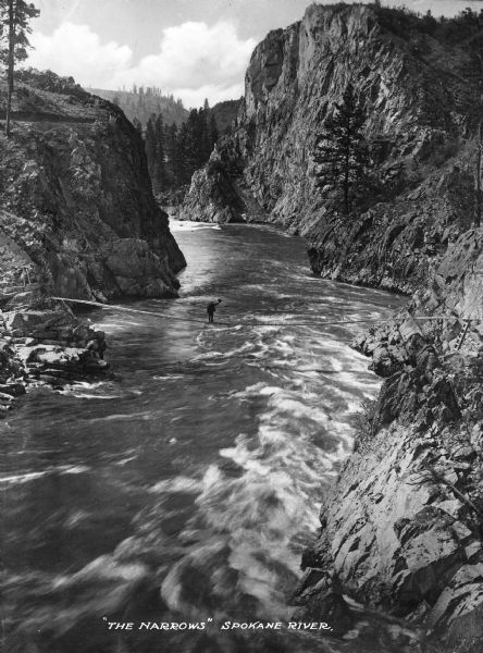 High, majestic view of "The Narrows" and surrounding cliffs of the Spokane River. A man is balancing on wires over the choppy water. Caption reads: "'The Narrows' Spokane River."